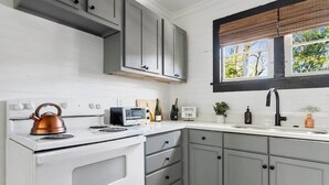 Una cocina con armarios blancos y grises, una estufa blanca, un hervidor de cobre, un tostador y una ventana con persianas.