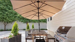 Un patio al aire libre con una mesa y sillas de madera, una parrilla y una sombrilla para dar sombra.