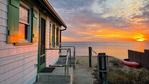A beachfront house with a concrete staircase leading to a porch, a kayak on the sand, and a sunset over the ocean. - Water Front Days Cottage #15 (2255) (North Truro)