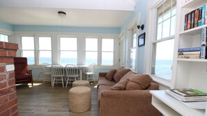 A living room with a brown sofa, a white bookshelf, and a view of the sea through large windows. - Water Front Days Cottage #15 (2255) (North Truro)