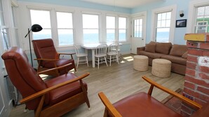 A room with a view of the sea, a dining table with chairs, a sofa, and a brick fireplace. - Water Front Days Cottage #15 (2255) (North Truro)