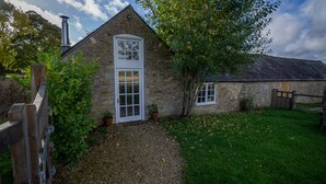 Une petite maison en pierre avec une porte et une fenêtre blanches, entourée d’un chemin de gravier et d’une pelouse verdoyante.