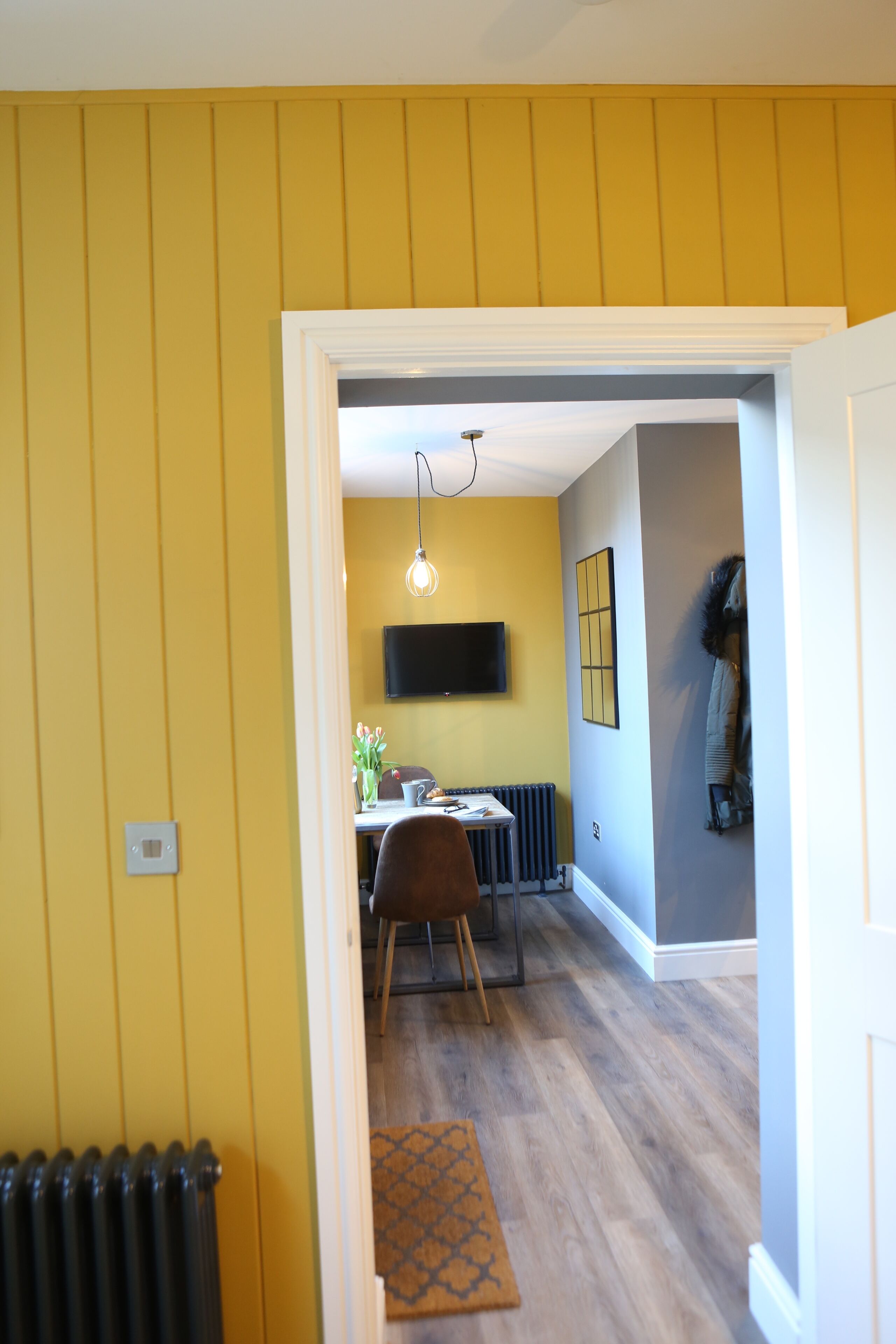 A hallway leading to a dining area with a table and chairs, a mounted TV, and a radiator on the wall.
