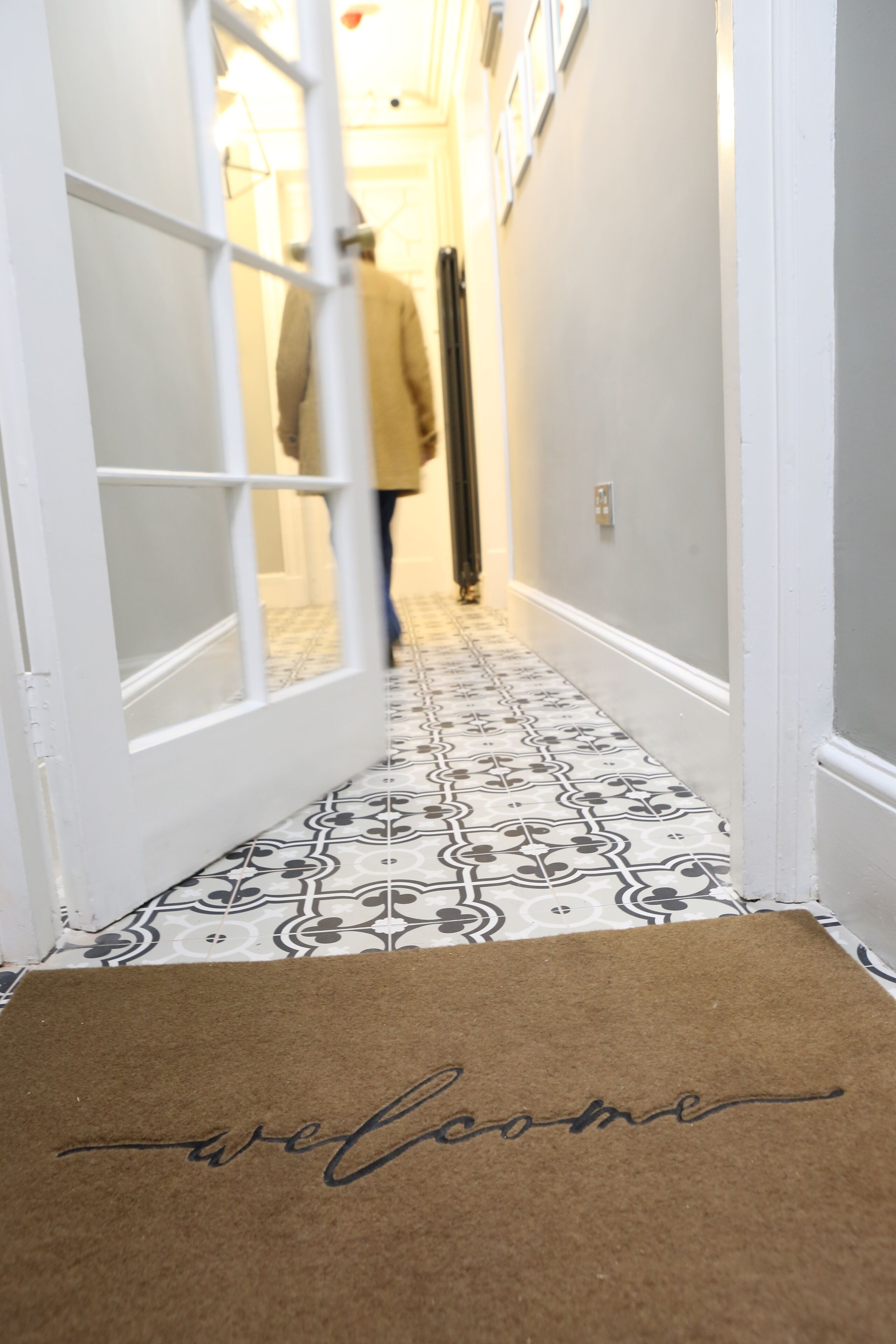 A hallway with patterned floor tiles, a welcome mat, and a person walking away.