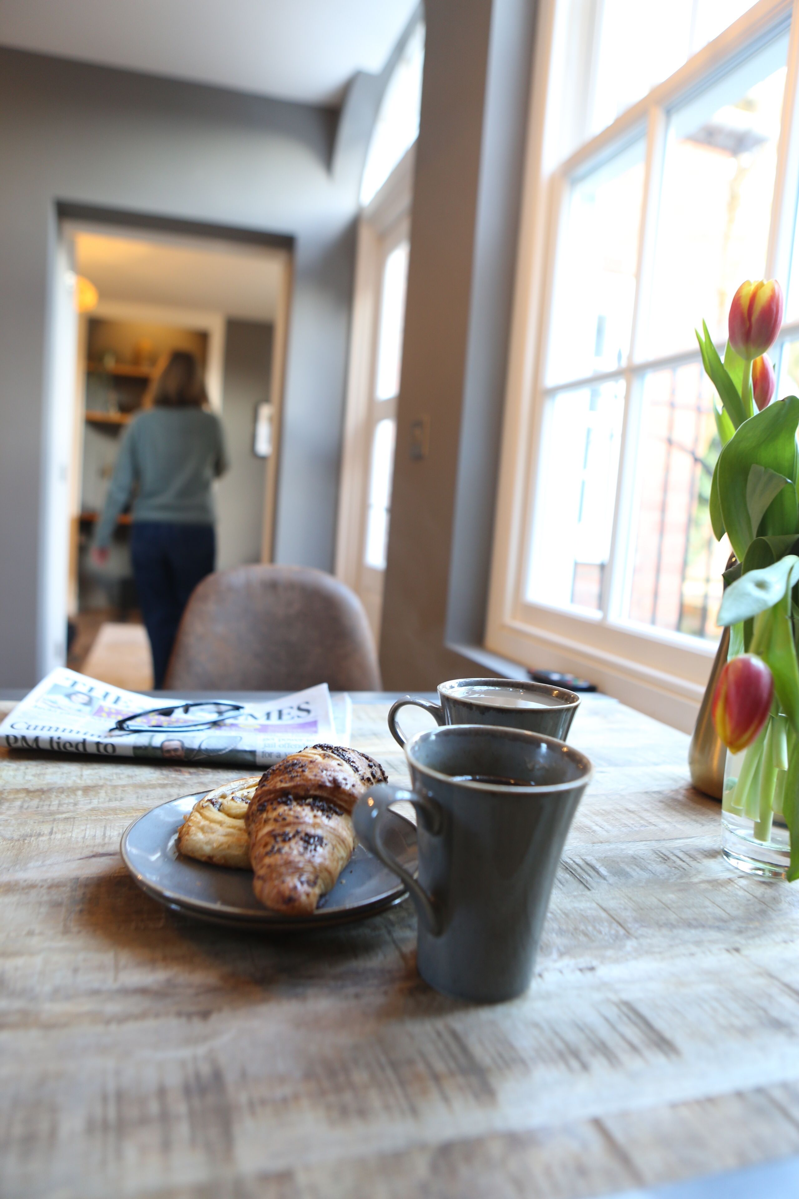 A breakfast table with croissants, a mug, and tulips by a window.