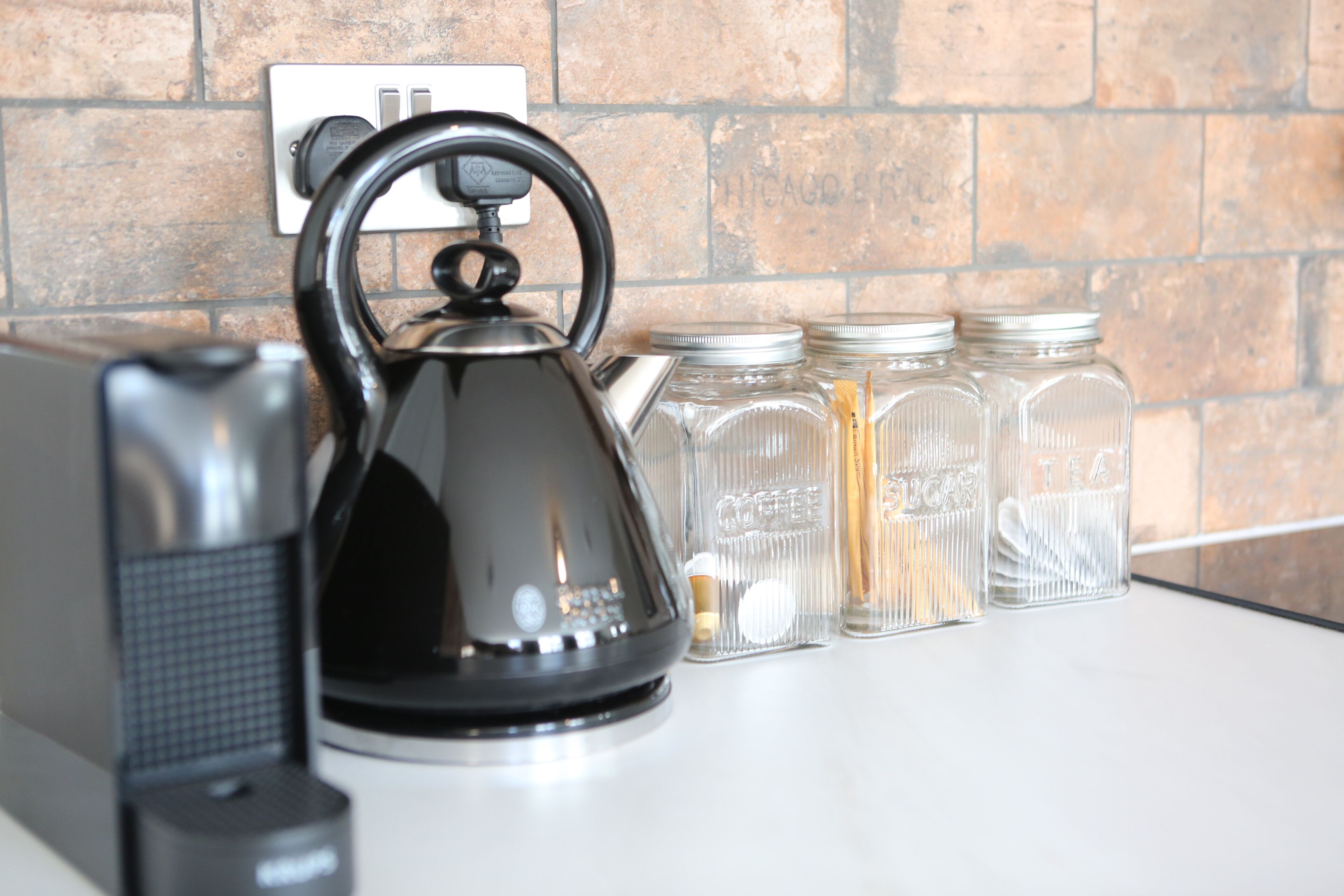 Un plan de travail de cuisine avec une bouilloire, une machine à café et trois bocaux en verre étiquetés avec des indications sur leur contenu.