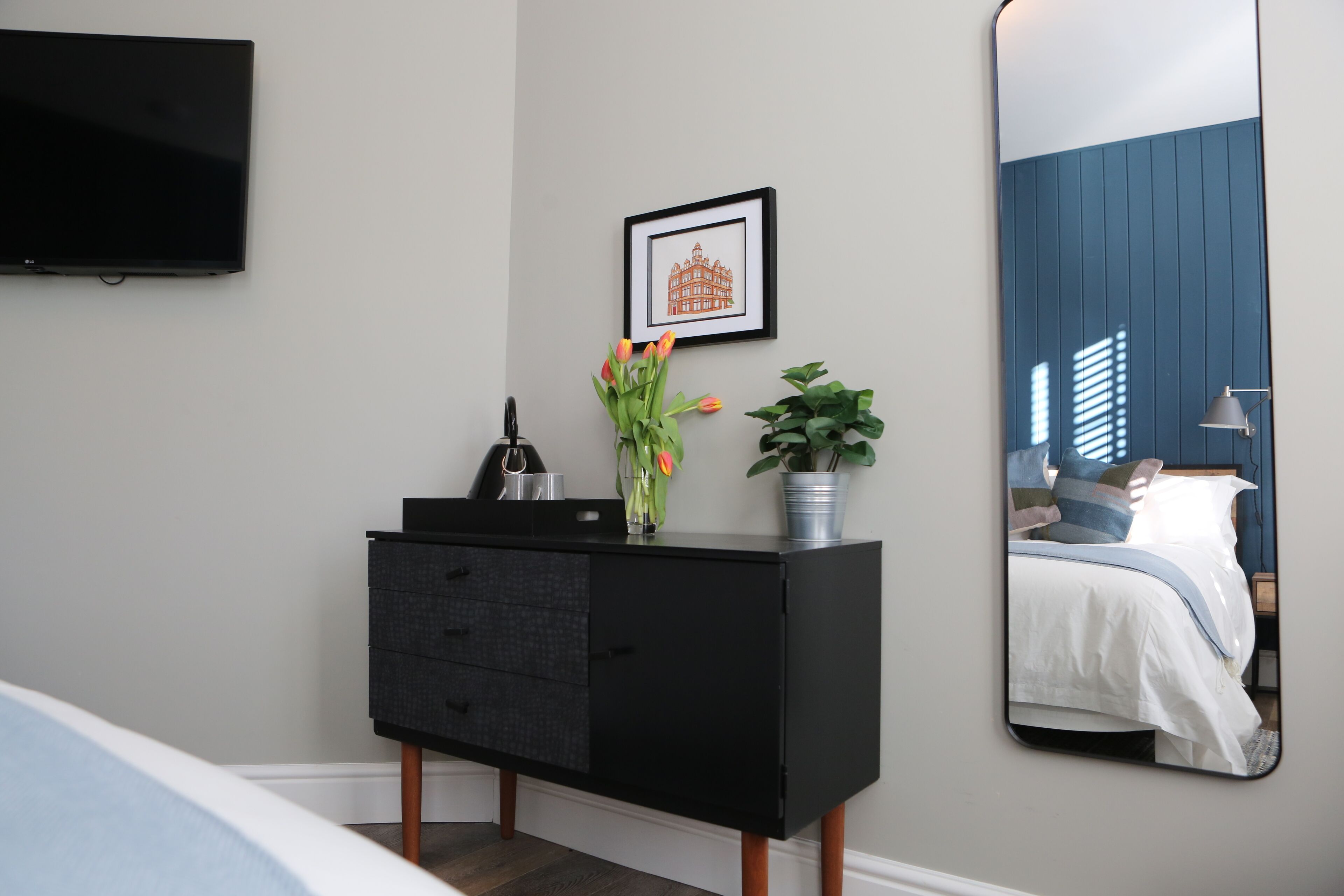A bedroom with a black dresser, a framed picture on the wall, a potted plant, and a large mirror reflecting the bed and window.