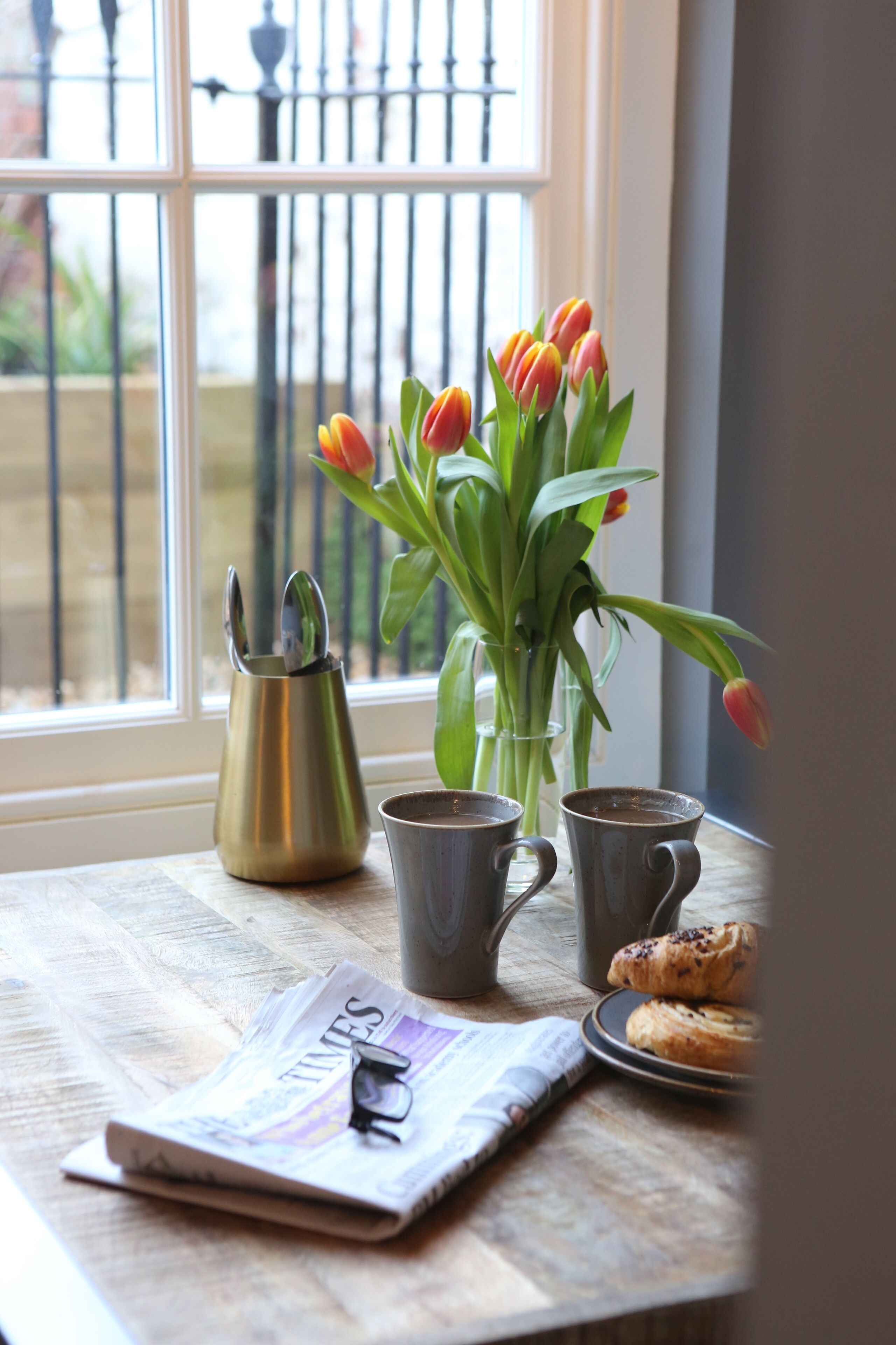 A table with a newspaper, a pair of glasses, a coffee pot, two mugs, and a plate with pastries.