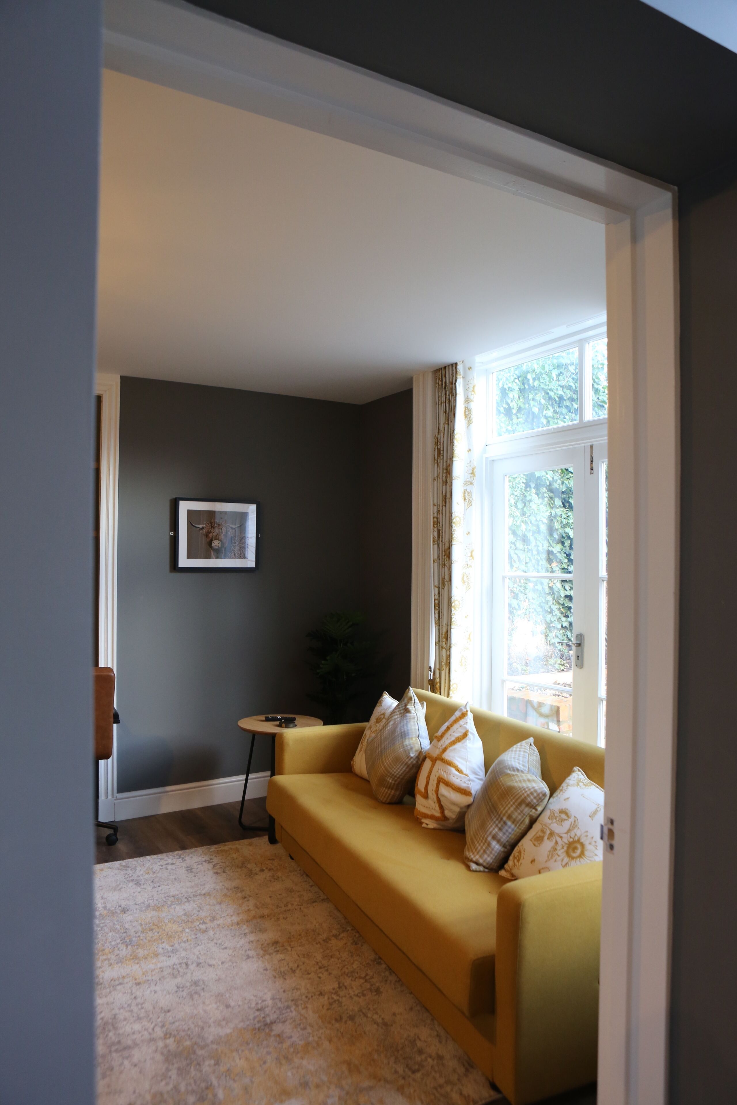 A living room with a yellow sofa, a framed picture on the wall, and a window with curtains.
