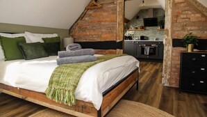 Un dormitorio con cama de madera, almohadas verdes y blancas, una manta verde y una cocina visible a través de un umbral abierto.