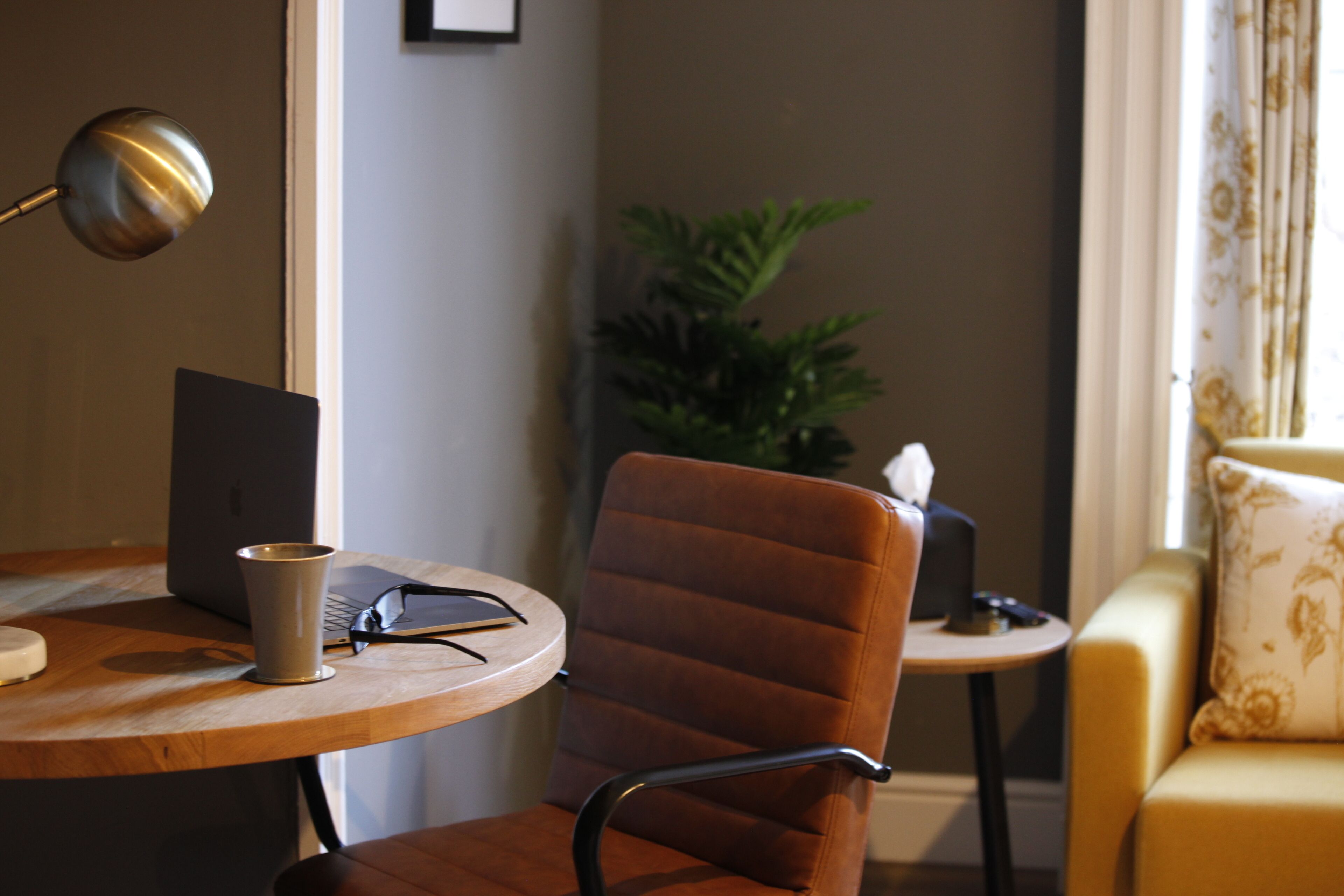 Une pièce avec un bureau en bois, un ordinateur portable, une tasse et une chaise. On y trouve une petite table avec une plante et une lampe. Un canapé avec un coussin est visible au fond de la pièce.