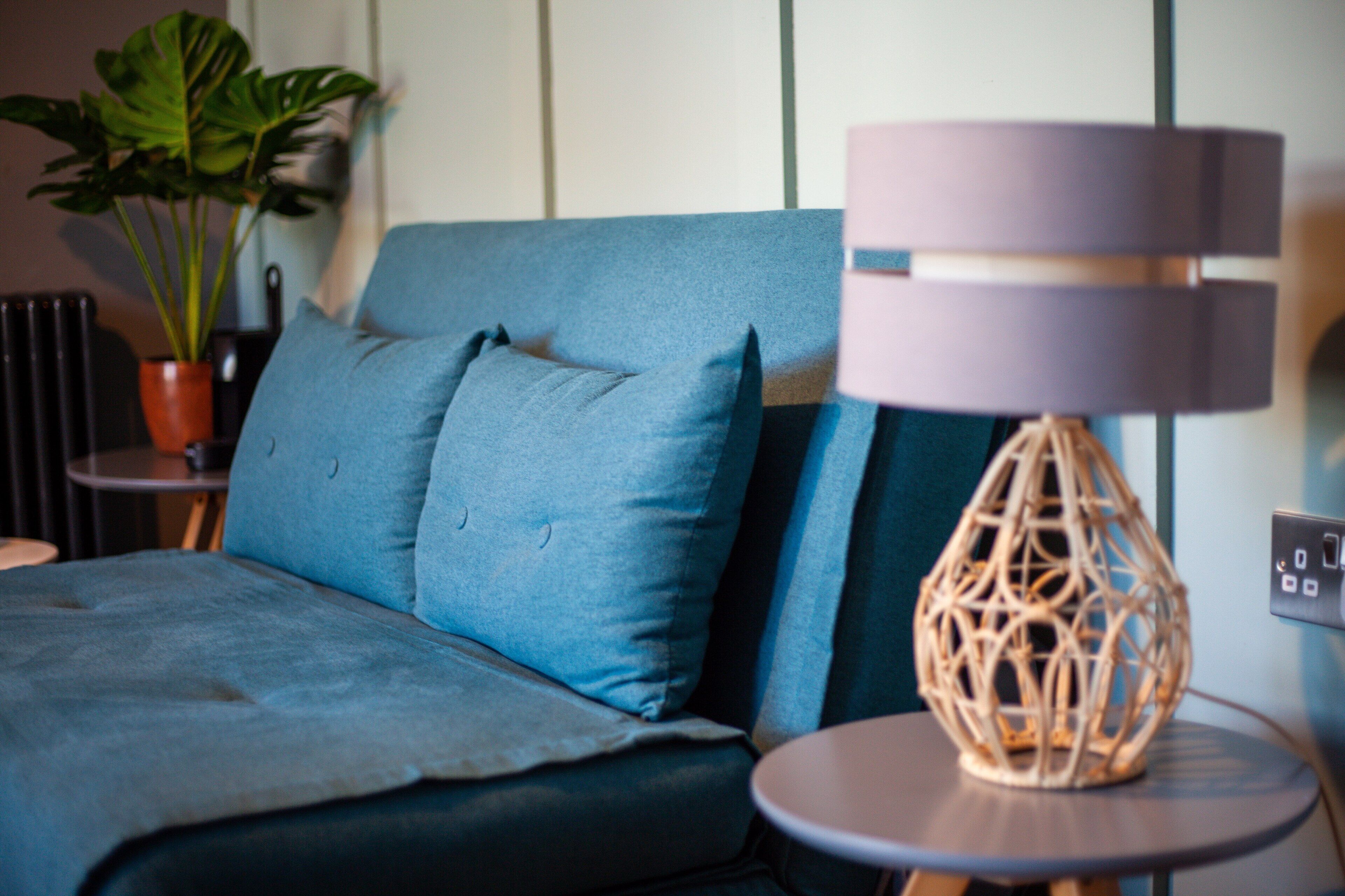 Et moderne soveværelse med en blå sofa, et rundt bord med en vævet lampe og en potteplante.