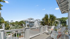 Terraza de madera con barandillas blancas y sillas, con vista a una zona residencial con casas y palmeras.