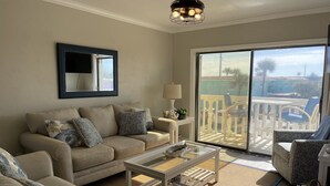 A living room with a beige sofa set, a coffee table, and a view of the outdoor balcony area. - Remodeled Beach Oasis (Ormond Beach)