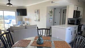 A modern living room with a glass dining table, brown woven placemats, a grey sofa, and a flat-screen TV. - Remodeled Beach Oasis (Ormond Beach)