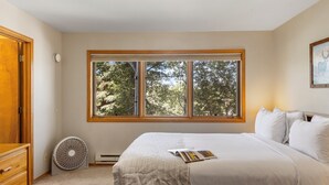 Une chambre avec une grande fenêtre, un lit avec du linge de lit blanc, un ventilateur et une porte en bois.