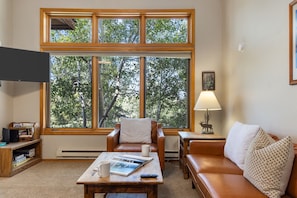 A cozy living room with a large window, a leather sofa, a coffee table with a book and mug, and a lamp.