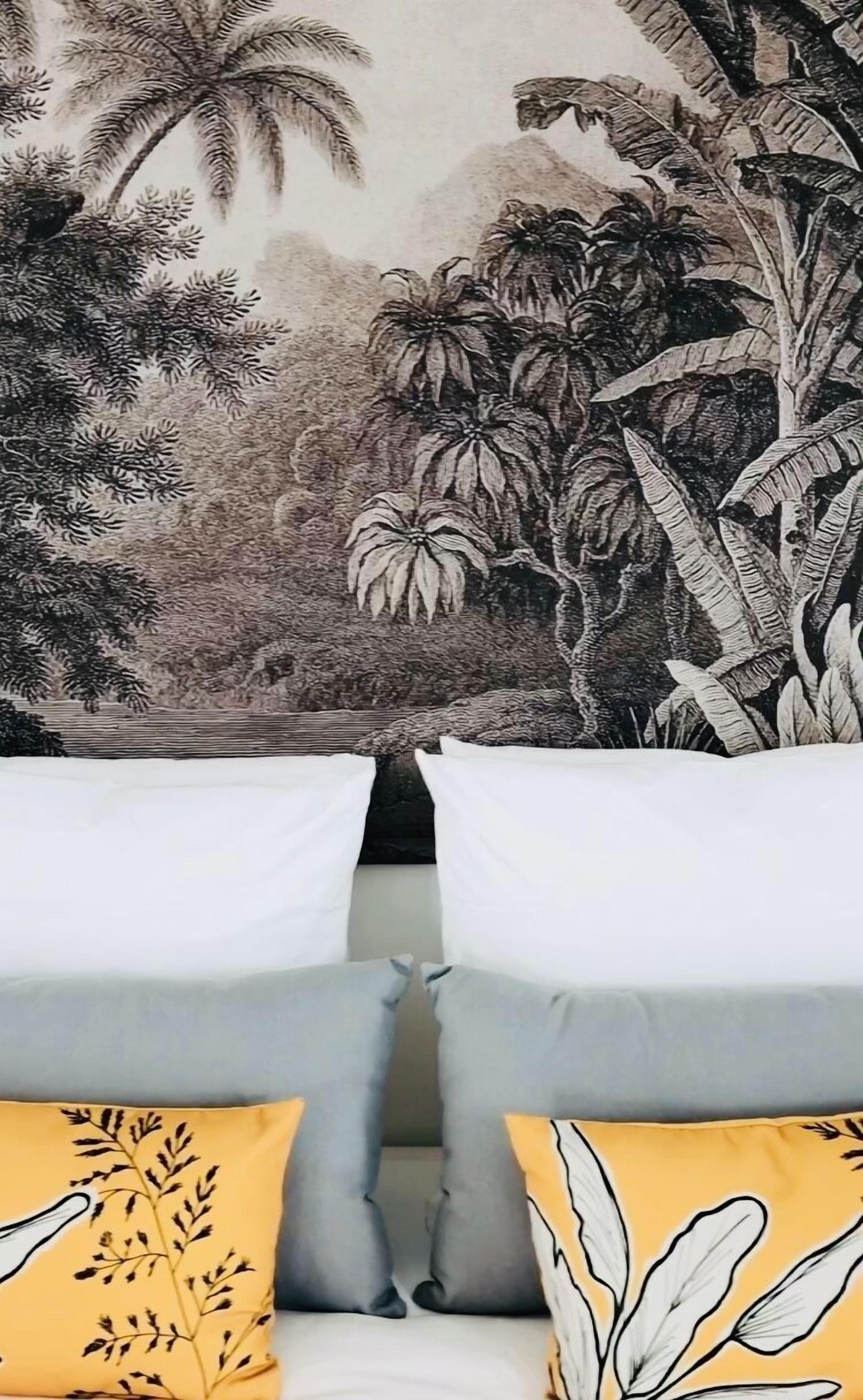 A bedroom with a large tropical mural on the wall, two white pillows, and a yellow pillow with a leaf pattern.