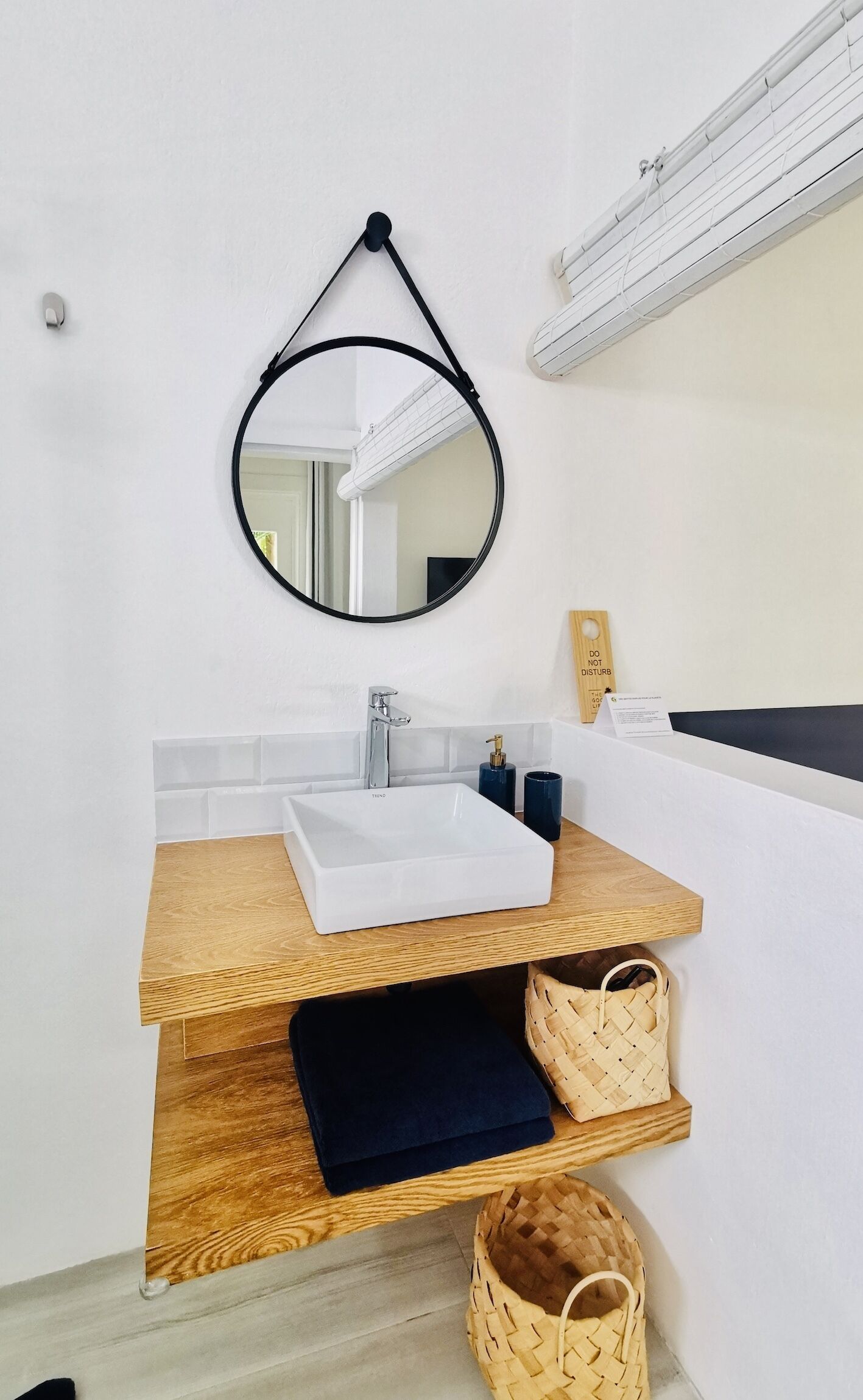 Un baño moderno con lavamanos blanco, tocador de madera, espejo redondo y perchero.
