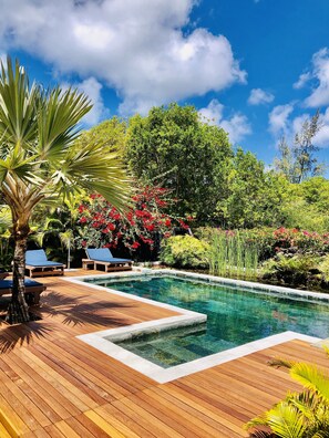 Una terraza de madera con piscina, rodeada de exuberante follaje y flores rojas.