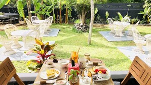 Un espacio de comedor al aire libre con mobiliario de mimbre blanco, una mesa dispuesta con platos de comida, un calentador de agua y un vaso de jugo.