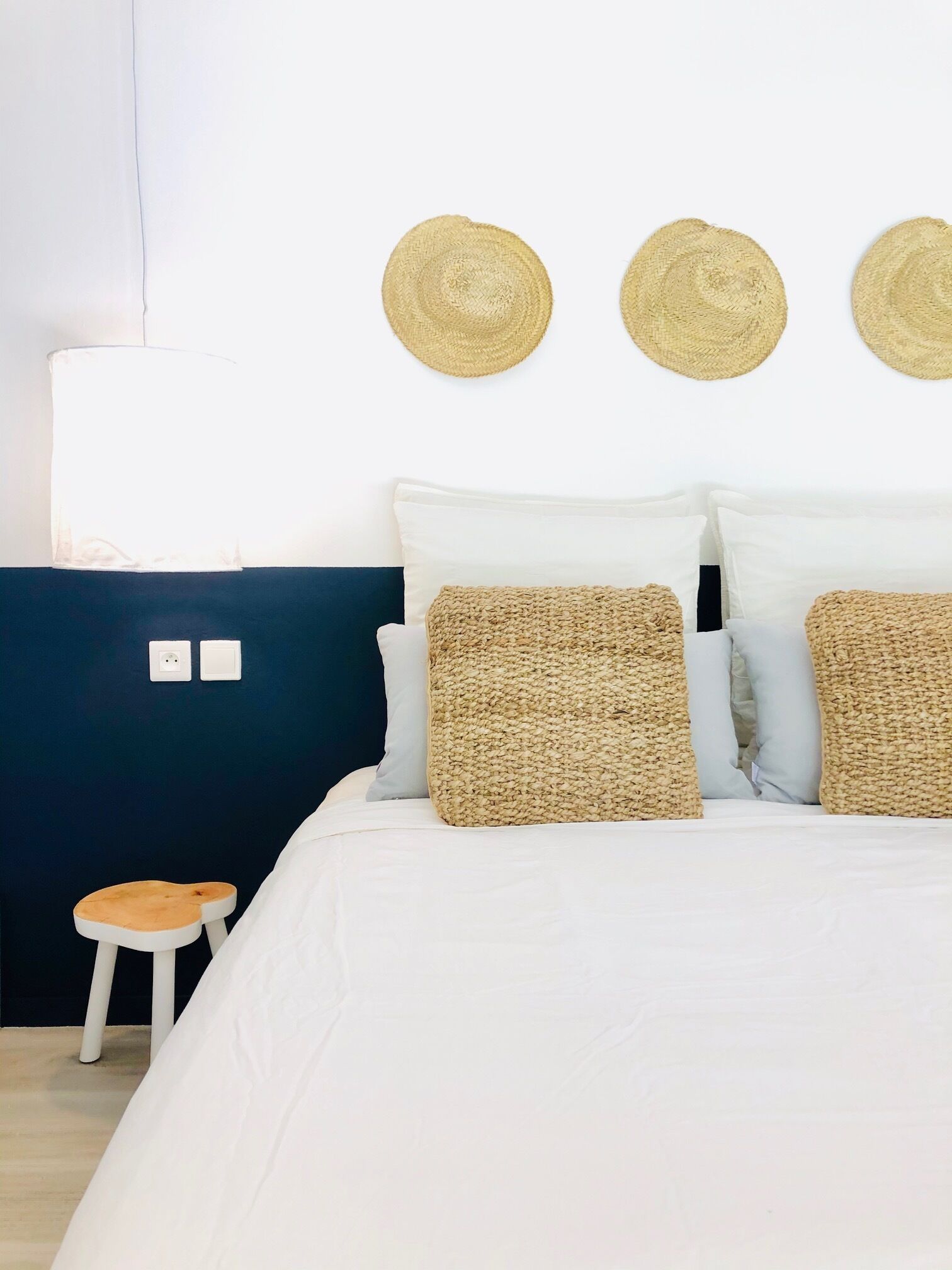 Una cama con ropa de cama blanca, dos almohadones tejidos y dos sombreros de paja en la pared.