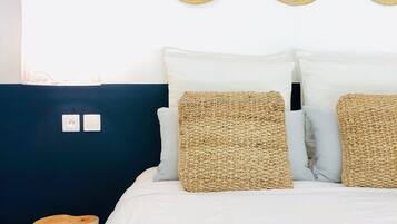 Una cama con ropa de cama blanca, dos almohadones tejidos y dos sombreros de paja en la pared.