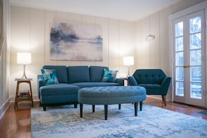 Ein Wohnzimmer mit Sofa, Hocker und Sessel, einem Gemälde an der Wand und einer Tür mit Blick.