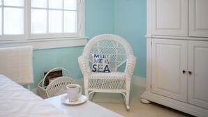 Um quarto com uma cadeira de vime, um armário branco e uma cesta com uma xícara sobre uma mesa.