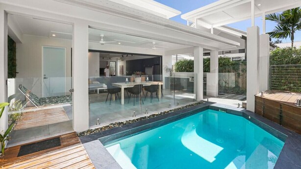Un área de piscina exterior moderna con un patio de cristal, una terraza de madera y una piscina de agua cristalina.