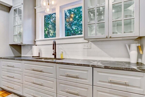 A kitchen with white cabinets, a granite countertop, and a window with a green leafy view.