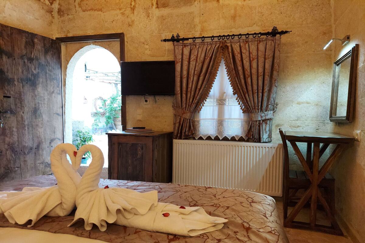A bedroom with a bed, a wooden chair, a small table, a window with curtains, and a view of the outside.