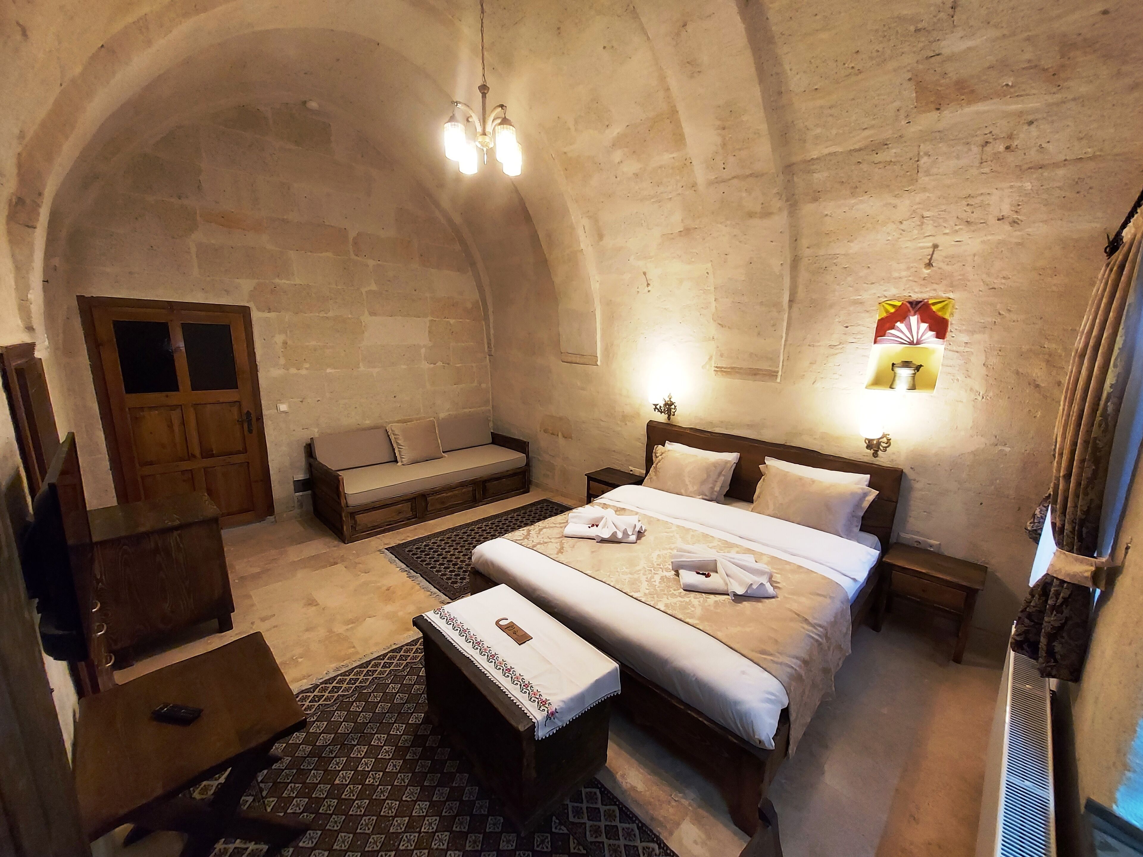Una habitación con pared de piedra, puerta de madera, cama con ropa blanca, banco de madera, una mesita y una lámpara fijada a la pared.