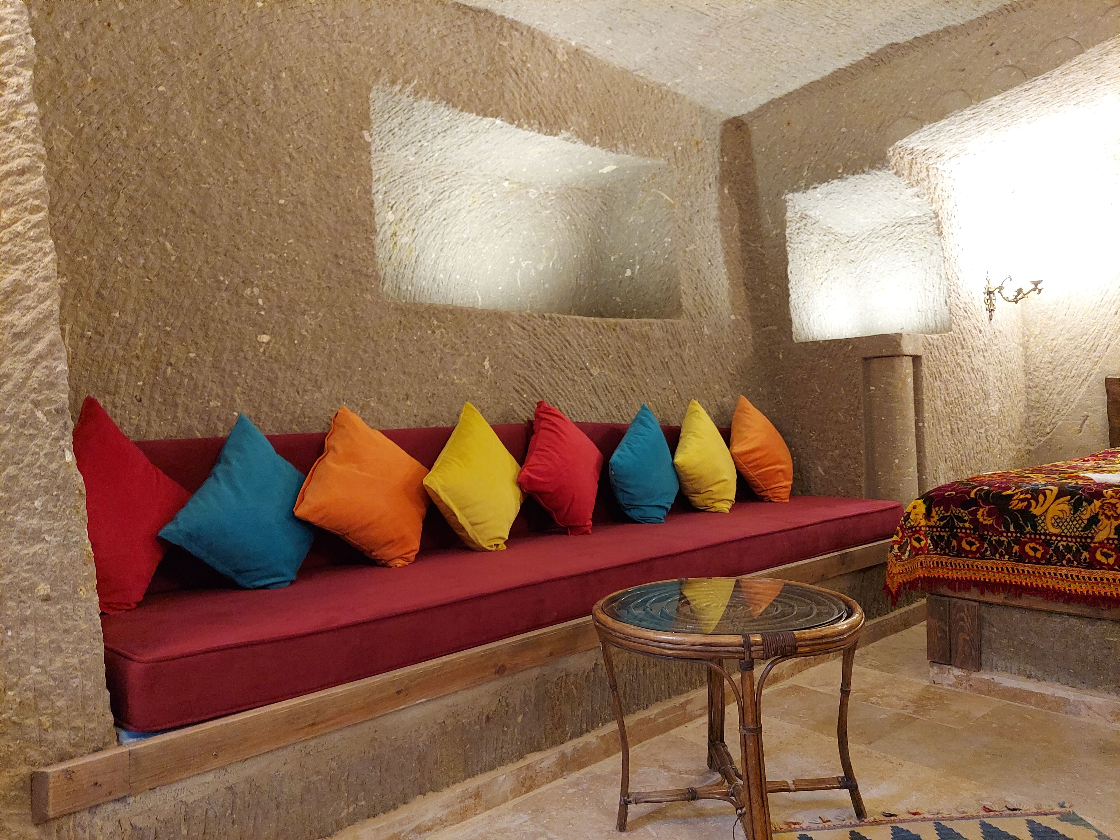 A room with a red sofa, colorful cushions, a glass table, and a patterned bedspread.
