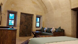 A rustic room with a wooden desk, a bed, a sofa, and two windows with wooden frames. - Armesos Cave Hotel (Ürgüp)