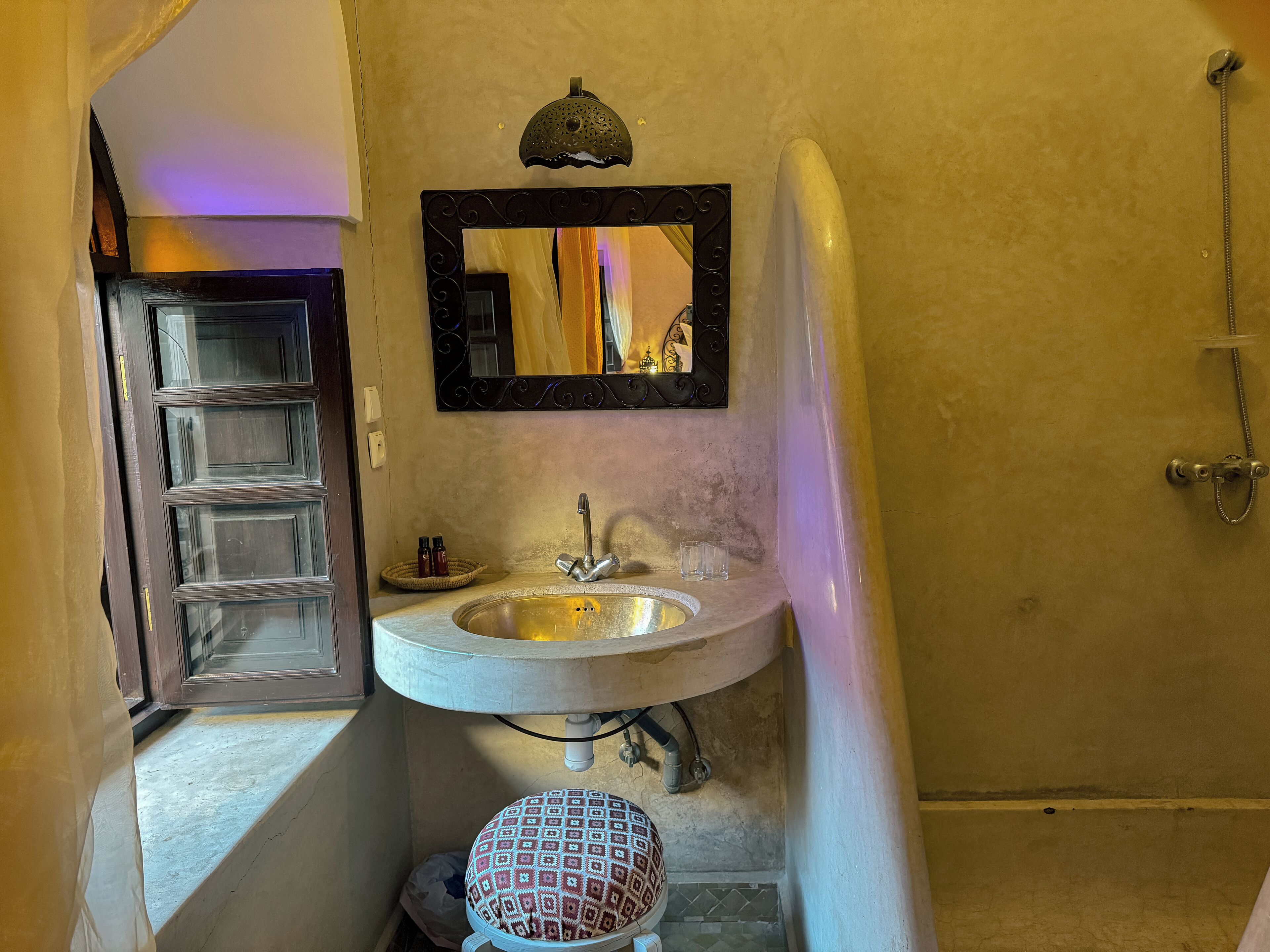 A bathroom with a sink, mirror, shower, and a patterned stool.
