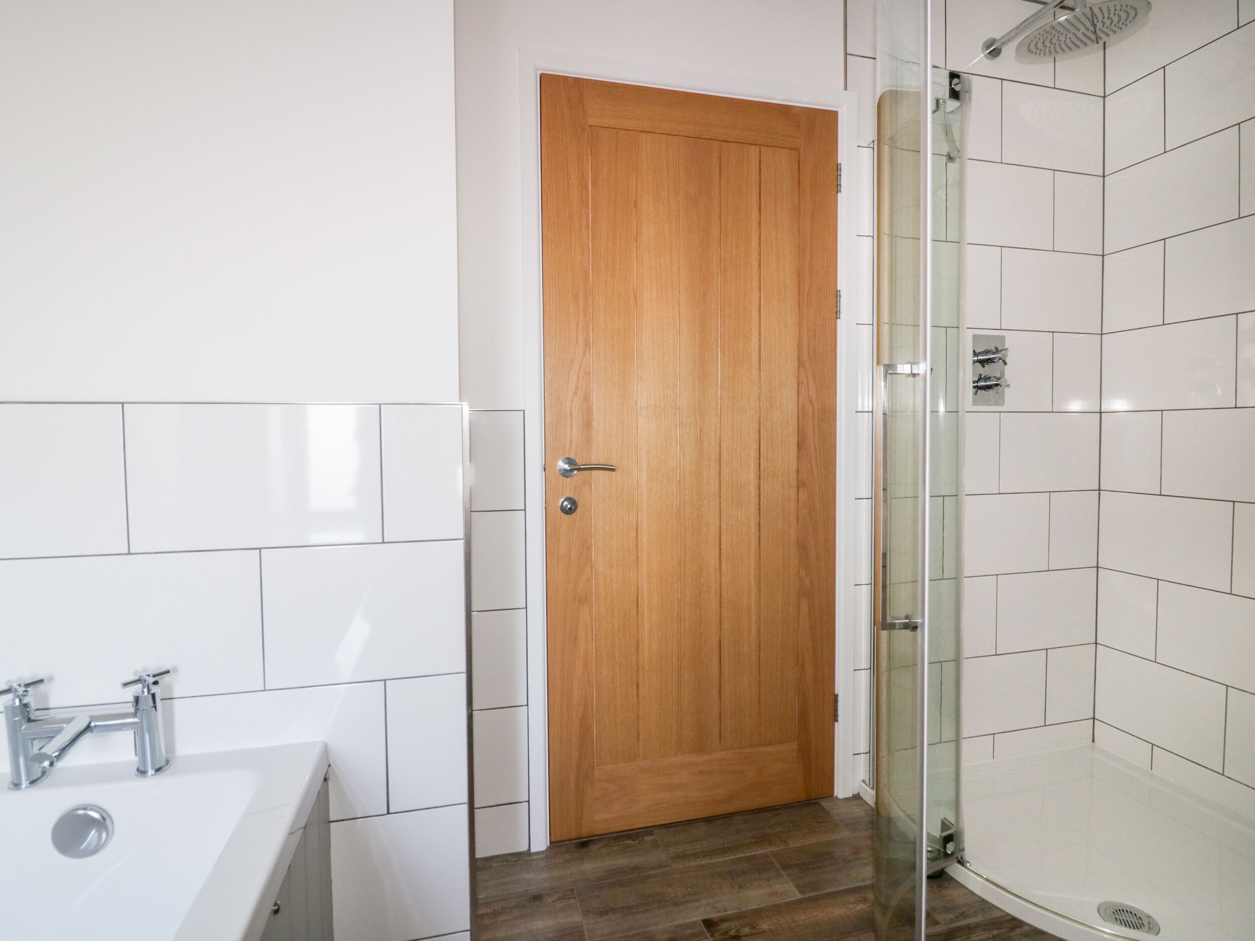 Een badkamer met een houten deur, een douche met witte tegels en een witte wastafel met chroombeslag.