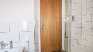 Een badkamer met een houten deur, een douche met witte tegels en een witte wastafel met chroombeslag.