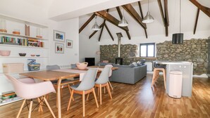 Een ruime woonkamer met een houten eettafel, stoelen en een grijze bank. Achtergrond toont een stenen muur en een open haard.