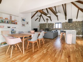 A spacious living area with a wooden dining table, chairs, and a grey sofa. A stone wall and a fireplace are visible in the background.