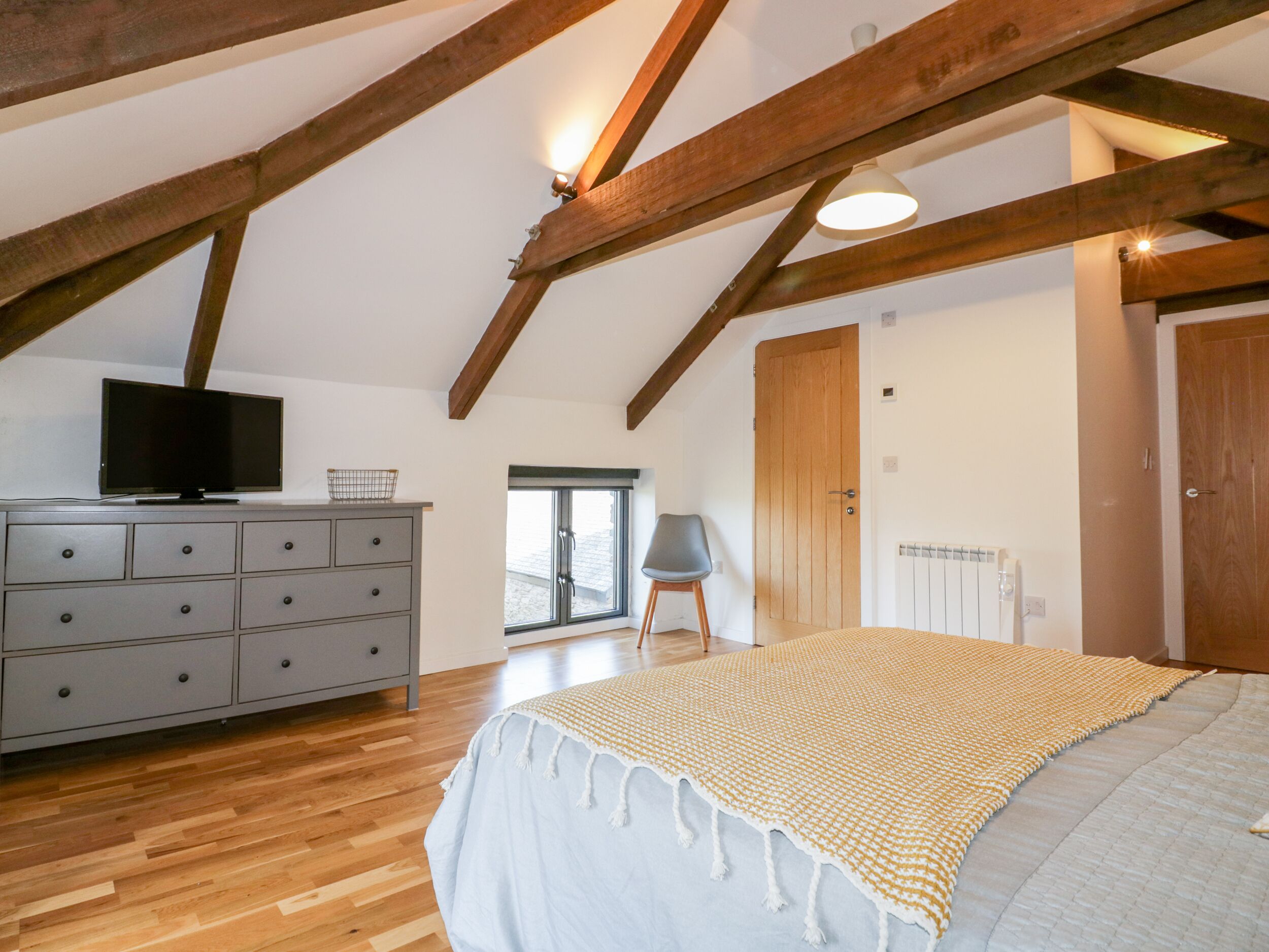 A bedroom with a wooden ceiling, a bed with a white and yellow blanket, a grey dresser, a television on top, and a window with a view.