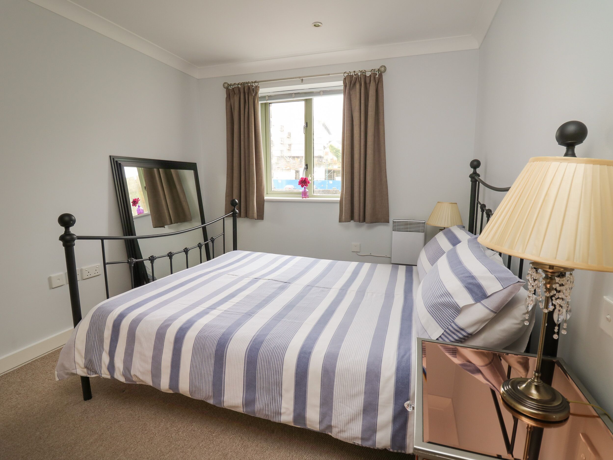 A neatly arranged bedroom with a bed, a bedside table with a lamp, a mirror, and a window with curtains.