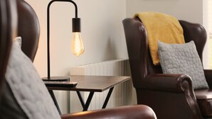 Una sala de estar acogedora con un sillón de cuero, una mesita de madera y una lámpara de pie con una bombilla visible.