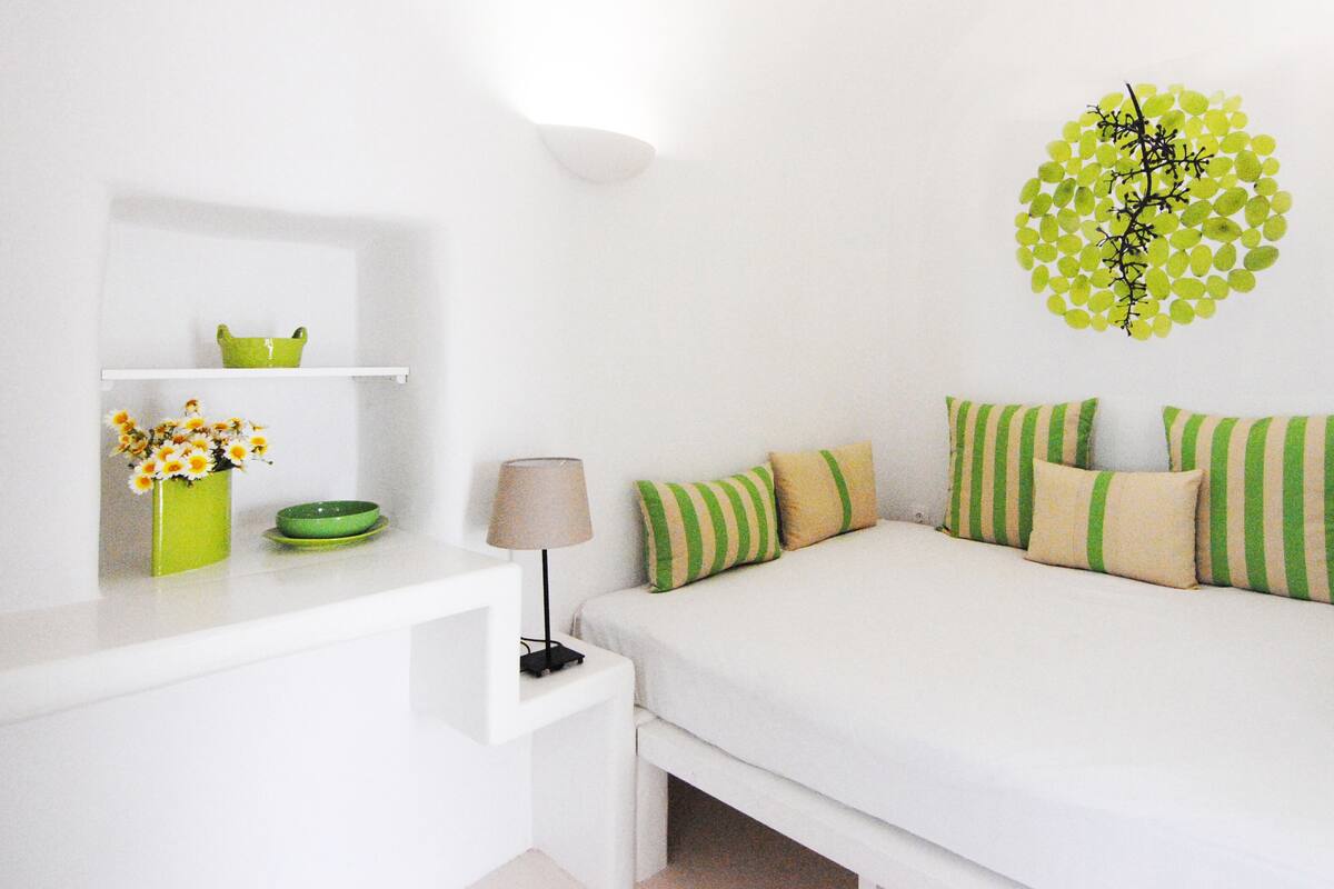 A white bedroom with a bed, green and beige striped pillows, a green vase with flowers, and a wall decoration.