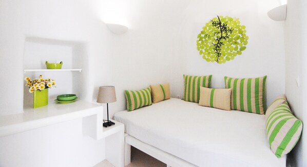 A white bedroom with a bed, green and beige striped pillows, a green vase with flowers, and a wall decoration. - White Grape Suites (Santorini)
