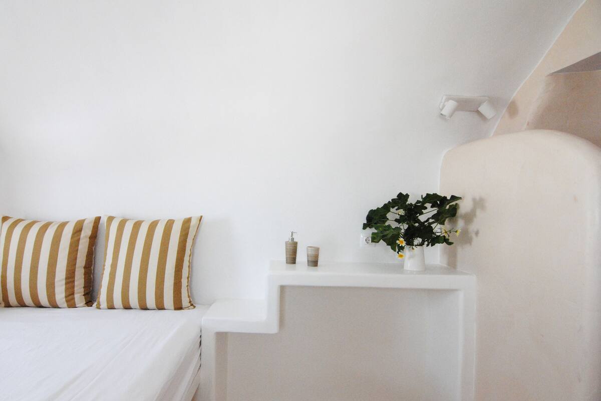 A bedroom with a bed, two striped pillows, a nightstand, a small vase with flowers, and a wall-mounted light fixture.