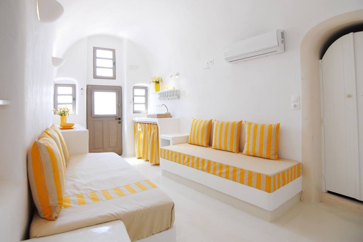 A bright, white room with a sofa and a bench, both with yellow and white striped cushions. There is a small table with a vase of flowers, and a window with curtains.