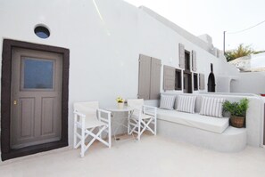 A white-walled building with a door, a small table with chairs, and a white sofa with pillows. - White Grape Suites (Santorini)