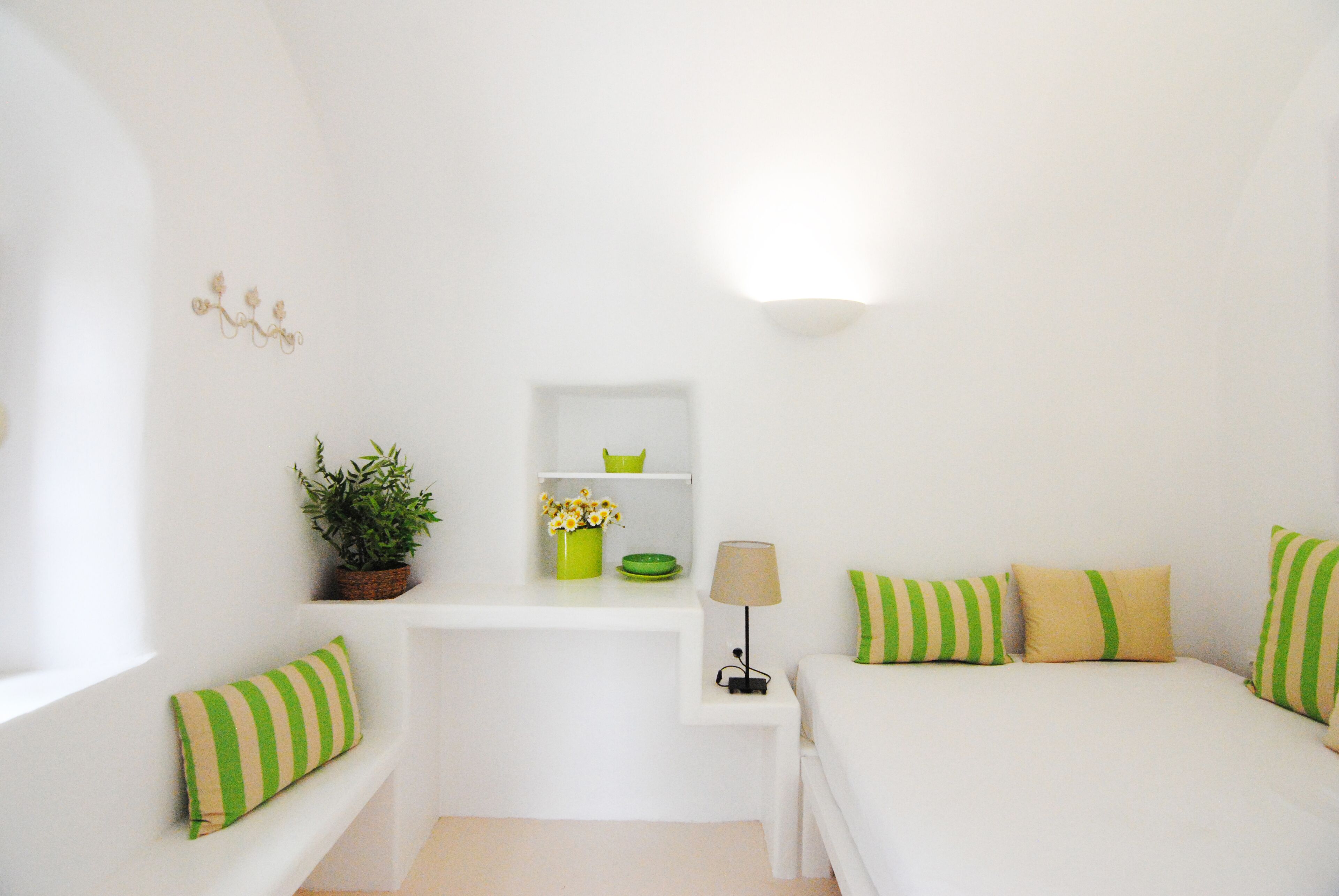 Une pièce blanche comprenant un lit, une table de chevet, une lampe, une étagère avec des objets décoratifs et une fenêtre donnant sur un paysage.