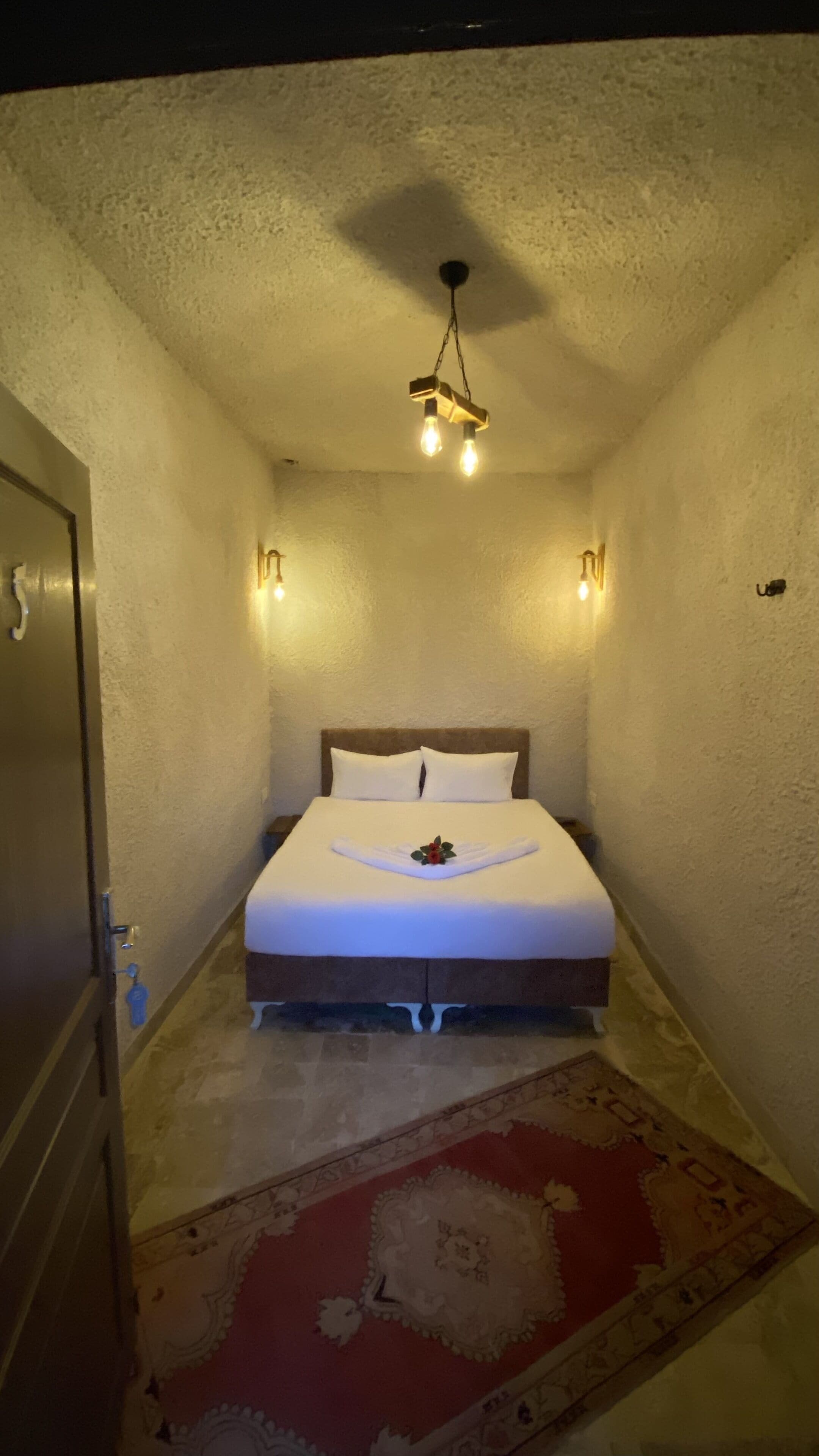 A narrow hotel room with a bed, a hanging light fixture, and a red patterned rug.